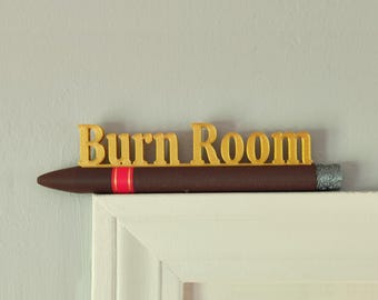 以下が含まれることがあります： 「Burn Room」と金色の文字で書かれた装飾的なサイン。茶色のボディ、赤いバンド、銀色の先端を持つ葉巻の形をしています。白い表面に置かれたサインです。