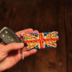 K&ouml;nnte beinhalten: Ein Schl&uuml;sselanh&auml;nger mit dem Text "RESTORE BRITAIN" in goldfarbenen Buchstaben, vor einem Union Jack-Design. Der Schl&uuml;sselanh&auml;nger ist an einem Autoschl&uuml;sselanh&auml;nger befestigt. Der Hintergrund ist eine dunkle Holzoberfl&auml;che.