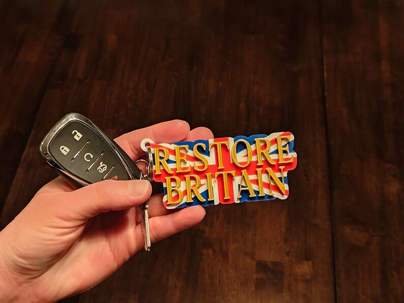 K&ouml;nnte beinhalten: Ein Schl&uuml;sselanh&auml;nger mit dem Text "RESTORE BRITAIN" in goldfarbenen Buchstaben, vor einem Union Jack-Design. Der Schl&uuml;sselanh&auml;nger ist an einem Autoschl&uuml;sselanh&auml;nger befestigt. Der Hintergrund ist eine dunkle Holzoberfl&auml;che.