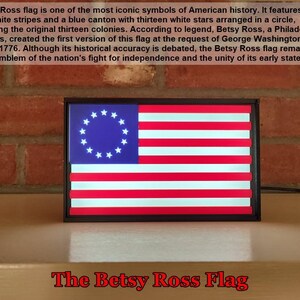 May include: A framed replica of the Betsy Ross flag, a historical American flag with thirteen red and white stripes and a blue canton with thirteen white stars arranged in a circle. The frame is black and the flag is illuminated. The text "The Betsy Ross Flag" is printed below the frame.