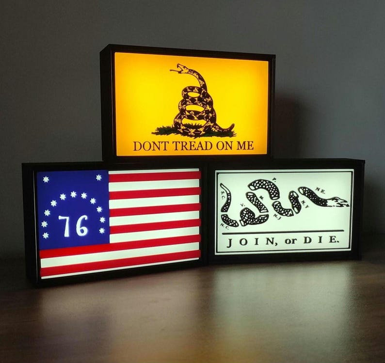May include: Three black framed light-up signs with white text and images. The top sign has a yellow background with a coiled snake and the text "DONT TREAD ON ME". The middle sign has a red, white, and blue American flag with 13 stars and the number "76" in the blue canton. The bottom sign has a white background with a coiled snake and the text "JOIN, or DIE."