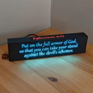 May include: A rectangular black light box featuring the red text Ephesians 6:11 and the blue illuminated text: Put on the full armour of God, so that you can take your stand against the devil's schemes. The device has a black power cord.