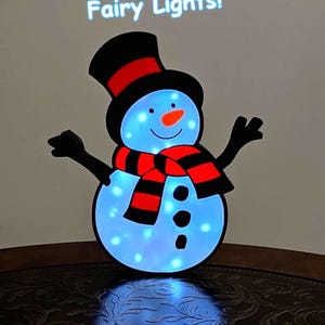 May include: A snowman-shaped decoration with blue fairy lights. The snowman has a black top hat and arms, a red and black scarf, and a carrot nose. The text "Fairy Lights!" is above the snowman.