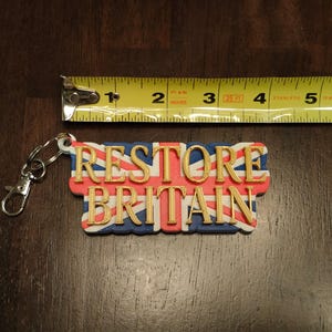 K&ouml;nnte beinhalten: Ein Schl&uuml;sselanh&auml;nger mit dem Text "RESTORE BRITAIN" in goldfarbenen Buchstaben, vor dem Hintergrund des Union Jack. Der Schl&uuml;sselanh&auml;nger ist ca. 7.6 cm lang und hat einen silbernen Verschluss.