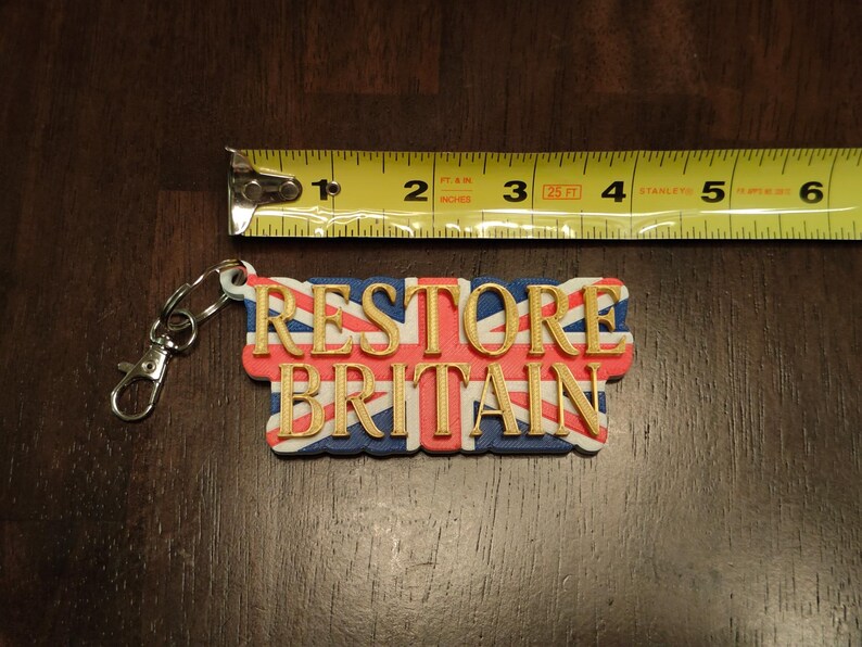 K&ouml;nnte beinhalten: Ein Schl&uuml;sselanh&auml;nger mit dem Text "RESTORE BRITAIN" in goldfarbenen Buchstaben, vor dem Hintergrund des Union Jack. Der Schl&uuml;sselanh&auml;nger ist ca. 7.6 cm lang und hat einen silbernen Verschluss.