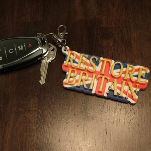 K&ouml;nnte beinhalten: Ein Satz Autoschl&uuml;ssel und ein Schl&uuml;sselanh&auml;nger. Der Schl&uuml;sselanh&auml;nger zeigt den Text "RESTORE BRITAIN" in goldfarbenen Buchstaben &uuml;ber einem Union Jack-Flaggen-Design. Die Schl&uuml;ssel sind schwarz und silber.