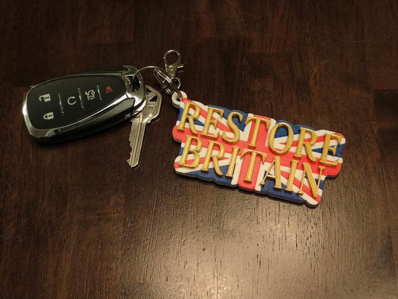K&ouml;nnte beinhalten: Ein Satz Autoschl&uuml;ssel und ein Schl&uuml;sselanh&auml;nger. Der Schl&uuml;sselanh&auml;nger zeigt den Text "RESTORE BRITAIN" in goldfarbenen Buchstaben &uuml;ber einem Union Jack-Flaggen-Design. Die Schl&uuml;ssel sind schwarz und silber.