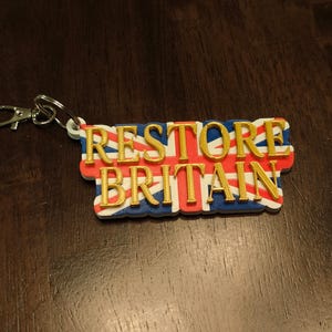 K&ouml;nnte beinhalten: Ein Schl&uuml;sselanh&auml;nger mit dem goldfarbenen Schriftzug "RESTORE BRITAIN". Der Hintergrund zeigt die britische Flagge, den Union Jack, in Rot, Wei&szlig; und Blau. Mit silberfarbenem Verschluss.