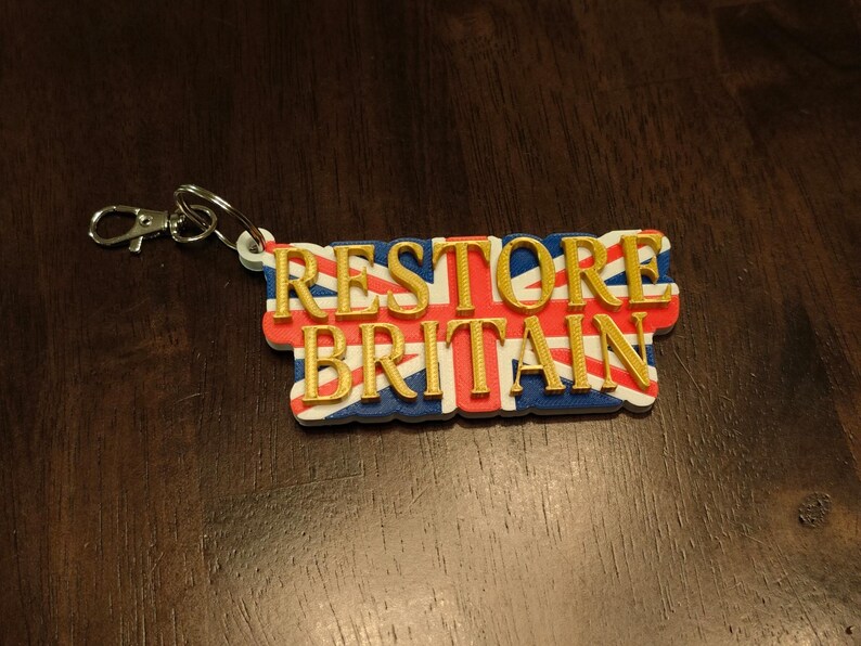K&ouml;nnte beinhalten: Ein Schl&uuml;sselanh&auml;nger mit dem goldfarbenen Schriftzug "RESTORE BRITAIN". Der Hintergrund zeigt die britische Flagge, den Union Jack, in Rot, Wei&szlig; und Blau. Mit silberfarbenem Verschluss.
