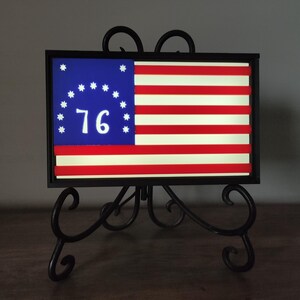 May include: A framed American flag with 13 stars in a circle and the number 76 in the center. The flag is displayed on a black metal stand.