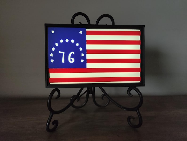 May include: A framed American flag with 13 stars in a circle and the number 76 in the center. The flag is displayed on a black metal stand.