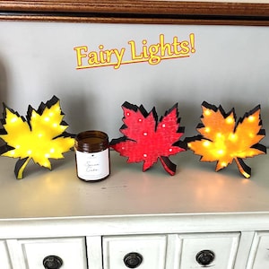 Pode incluir: Três decorações de folhas de bordo iluminadas em amarelo, vermelho e laranja, com bordas pretas. Um frasco de vela perfumada fica entre as folhas. O texto "Fairy Lights!" está acima das folhas.