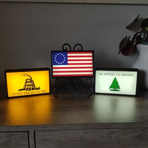 May include: Three illuminated framed prints with historical American flag designs. The first print features a yellow background with a coiled snake and the text "Don't Tread On Me". The second print features a red, white, and blue American flag with 13 stars in a circle. The third print features a white background with a green pine tree and the text "An Appeal To Heaven".