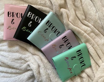 May include: Five colorful faux leather wallets in pink, black, mint, lavender, and green, each with the words "BROKE & Boujee" printed on the front. The wallets are arranged on a soft, white, textured surface.