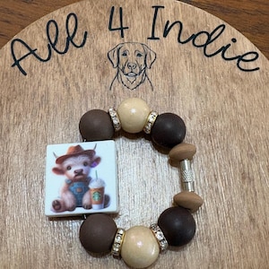 May include: A handmade bracelet with a square charm featuring a cartoon calf in a cowboy hat holding a coffee cup. The bracelet has brown, tan, and silver beads. The wooden background has the text "All 4 Indie" and a dog illustration.