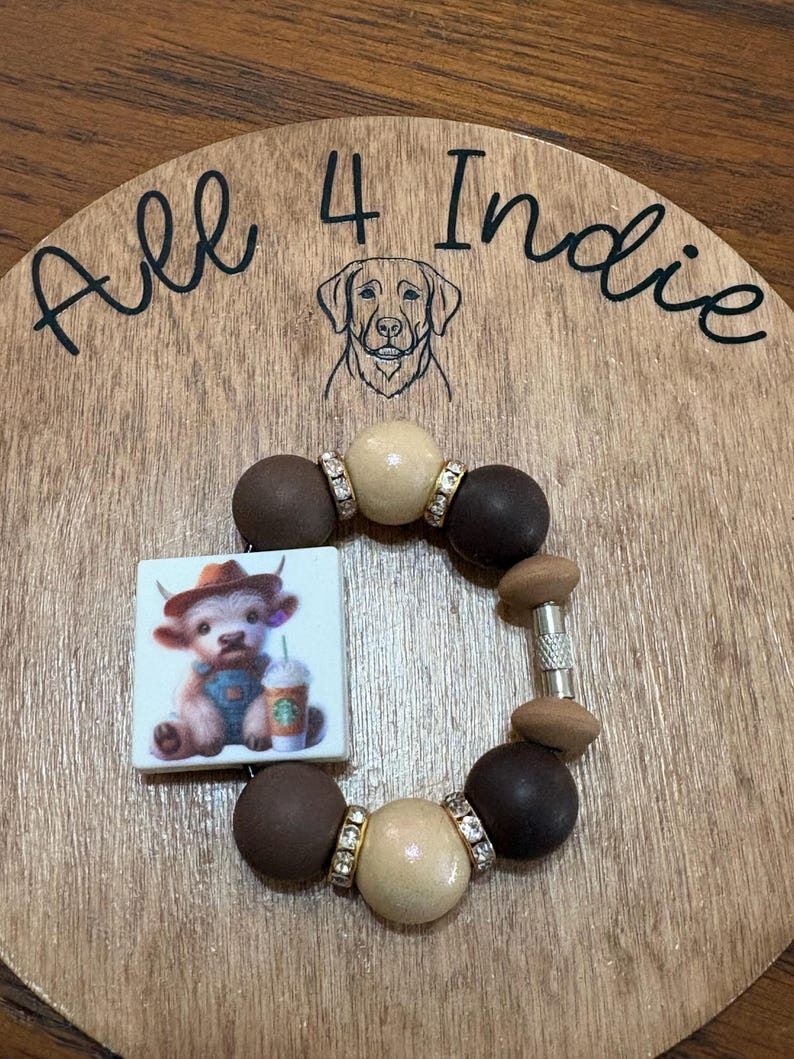 May include: A handmade bracelet with a square charm featuring a cartoon calf in a cowboy hat holding a coffee cup. The bracelet has brown, tan, and silver beads. The wooden background has the text "All 4 Indie" and a dog illustration.