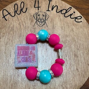 May include: A handmade bracelet with large pink and turquoise beads, accented with silver-toned spacers. A square charm with glitter and text is attached. The bracelet rests on a wooden surface with the text "All 4 Indie" and a dog illustration.
