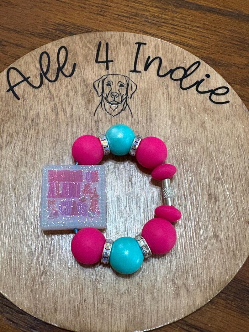 May include: A handmade bracelet with large pink and turquoise beads, accented with silver-toned spacers. A square charm with glitter and text is attached. The bracelet rests on a wooden surface with the text "All 4 Indie" and a dog illustration.