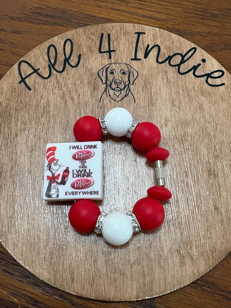 May include: A handmade bracelet with red and white beads, accented with silver-colored spacers. A small rectangular charm features the Dr. Pepper logo and the phrase "I WILL DRINK Dr. Pepper EVERYWHERE". The bracelet is displayed on a wooden surface with the text "All 4 Indie".
