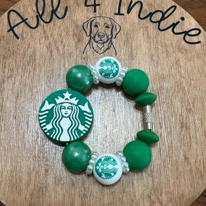 May include: A green and white beaded bracelet featuring the Starbucks logo. The bracelet includes green beads and white beads with the Starbucks logo. The bracelet is displayed on a wooden surface with the text "All 4 Indie" and a dog illustration.