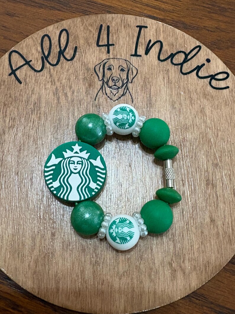 May include: A green and white beaded bracelet featuring the Starbucks logo. The bracelet includes green beads and white beads with the Starbucks logo. The bracelet is displayed on a wooden surface with the text "All 4 Indie" and a dog illustration.