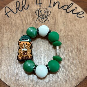 May include: A handmade bracelet featuring green and white beads, accented with black rhinestones. A charm shaped like a bear wearing a hat and holding a drink is attached. The bracelet is displayed on a wooden surface with the text "All 4 Indie".