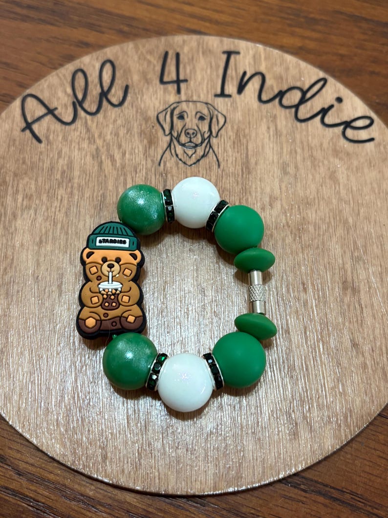 May include: A handmade bracelet featuring green and white beads, accented with black rhinestones. A charm shaped like a bear wearing a hat and holding a drink is attached. The bracelet is displayed on a wooden surface with the text "All 4 Indie".