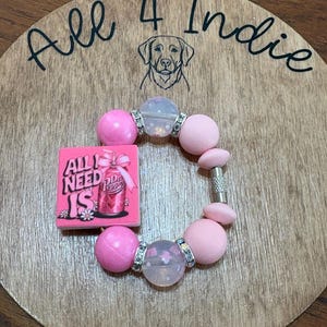May include: A pink beaded bracelet with a charm that reads "All I Need Is Dr. Pepper" on a wooden background. The bracelet features large pink beads, clear beads with silver accents, and a silver clasp. The background has the text "All 4 Indie" and a dog illustration.