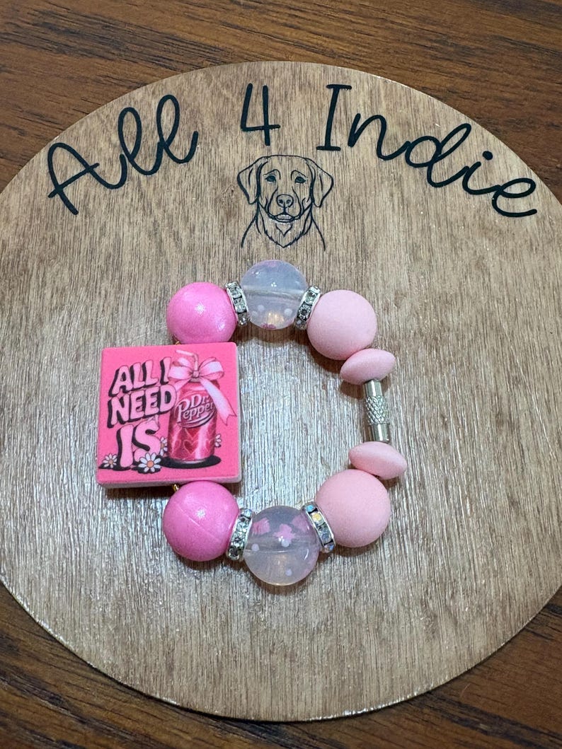 May include: A pink beaded bracelet with a charm that reads "All I Need Is Dr. Pepper" on a wooden background. The bracelet features large pink beads, clear beads with silver accents, and a silver clasp. The background has the text "All 4 Indie" and a dog illustration.