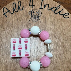 May include: A handmade bracelet featuring pink glitter beads, white beads, and a square charm with cherry designs. The bracelet includes rhinestone accents and a silver-tone clasp. The wooden background has the text "All 4 Indie" and a dog illustration.