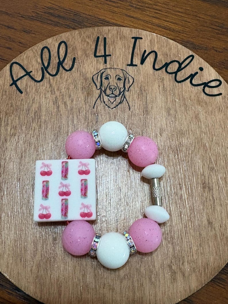 May include: A handmade bracelet featuring pink glitter beads, white beads, and a square charm with cherry designs. The bracelet includes rhinestone accents and a silver-tone clasp. The wooden background has the text "All 4 Indie" and a dog illustration.