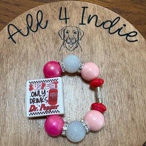 May include: A handmade bracelet featuring pink, light blue, and red beads. A square charm with the text "SUSPECT ONLY DRINKS Dr. Pepper" is included. The bracelet is set on a wooden background with the text "All 4 Indie".