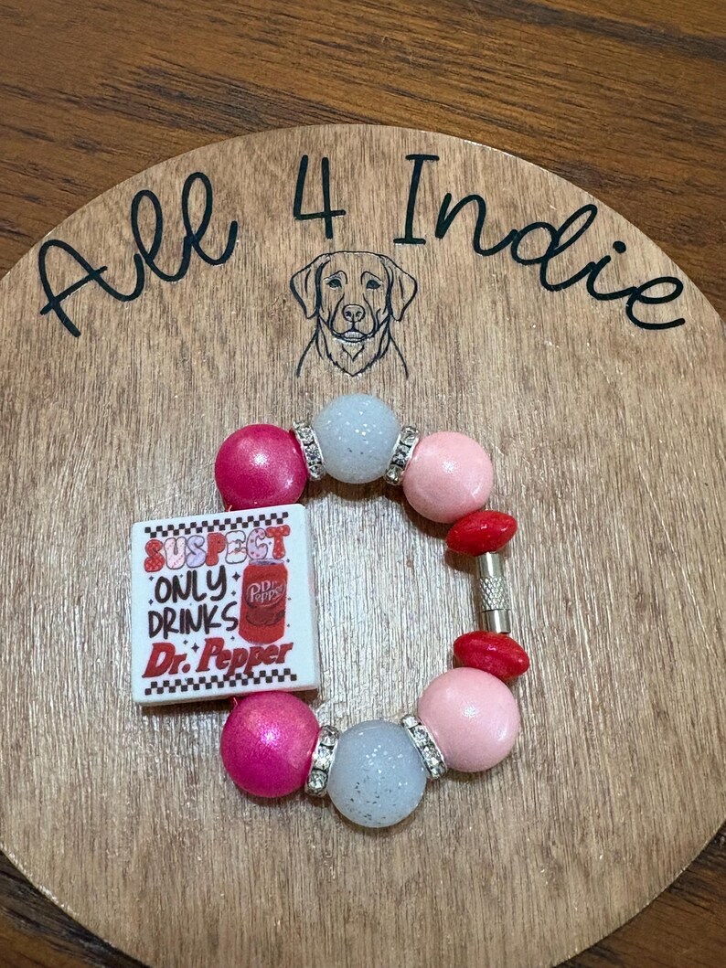 May include: A handmade bracelet featuring pink, light blue, and red beads. A square charm with the text "SUSPECT ONLY DRINKS Dr. Pepper" is included. The bracelet is set on a wooden background with the text "All 4 Indie".