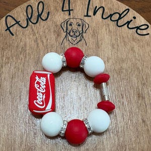 May include: A handmade bracelet featuring red and white beads, accented with silver-toned spacers. A miniature Coca-Cola can charm is attached. The bracelet is displayed on a wooden surface with the text "All 4 Indie" and a dog illustration.