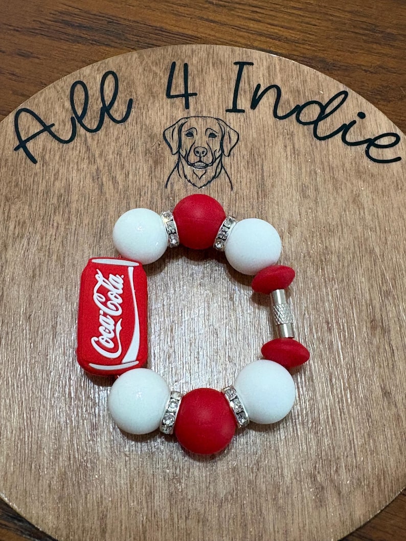 May include: A handmade bracelet featuring red and white beads, accented with silver-toned spacers. A miniature Coca-Cola can charm is attached. The bracelet is displayed on a wooden surface with the text "All 4 Indie" and a dog illustration.