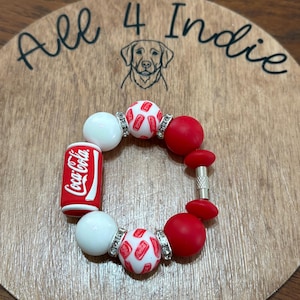 May include: A red, white, and silver beaded bracelet with a Coca-Cola can charm. The bracelet includes white and red round beads, and beads with a Coca-Cola logo. The bracelet is displayed on a wooden surface with the text "All 4 Indie".