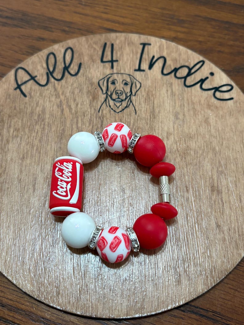 May include: A red, white, and silver beaded bracelet with a Coca-Cola can charm. The bracelet includes white and red round beads, and beads with a Coca-Cola logo. The bracelet is displayed on a wooden surface with the text "All 4 Indie".