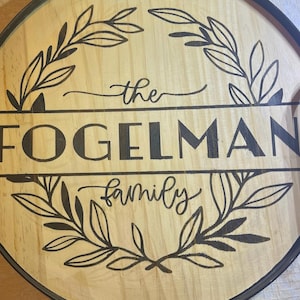 Puede incluir: Letrero redondo de madera con el texto "the FOGELMAN family" en letras negras. El letrero está rodeado por un marco de metal negro y presenta un diseño de hojas. El fondo es de madera clara, unos 30 cm de diámetro.
