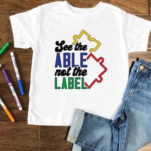 Może przedstawiać: Biała koszulka z kolorowym wzorem puzzli i tekstem "See the ABLE not the LABEL".