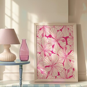 May include: A framed print of a pink floral design with white outlines. The print is hanging on a light beige wall. A pink glass vase and a lamp with a pink lampshade are on a blue table in front of the print.