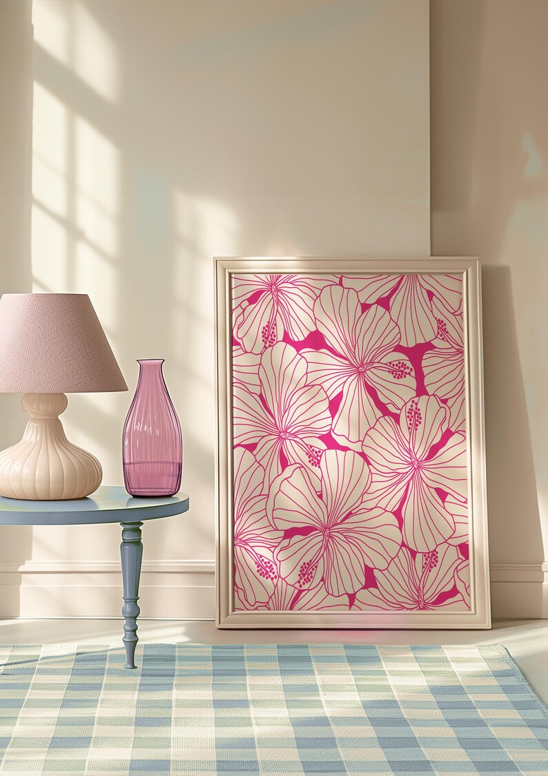 May include: A framed print of a pink floral design with white outlines. The print is hanging on a light beige wall. A pink glass vase and a lamp with a pink lampshade are on a blue table in front of the print.