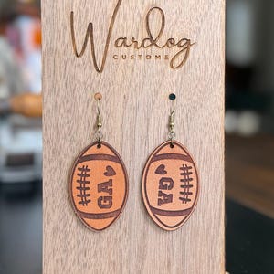 Op de afbeelding: Een paar oorbellen in de vorm van een voetbal met de letters "GA" en gestikte details. De oorbellen worden getoond op een houten standaard met het logo "Wardog Customs". De oorbellen zijn lichtbruin.
