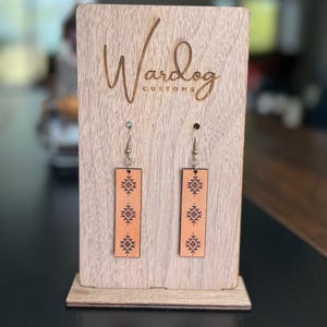 May include: A pair of rectangular, light brown leather earrings with a geometric pattern, displayed on a wooden stand. The stand is engraved with the text "Wardog CUSTOMS". The earrings hang from silver-colored hooks.