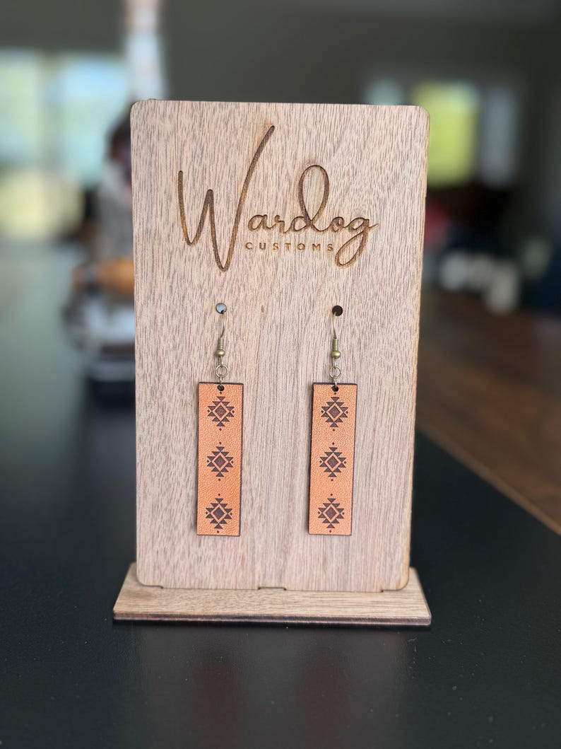 May include: A pair of rectangular, light brown leather earrings with a geometric pattern, displayed on a wooden stand. The stand is engraved with the text "Wardog CUSTOMS". The earrings hang from silver-colored hooks.