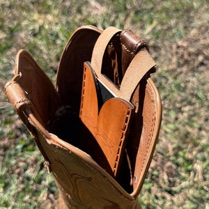 May include: A tan leather cowboy boot with decorative stitching. Inside the boot is a brown leather card holder with a black accent and a leather strap. The boot is set against a blurred green background.