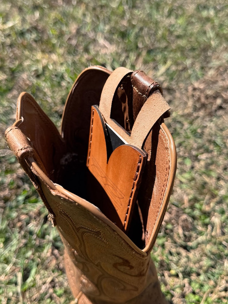 May include: A tan leather cowboy boot with decorative stitching. Inside the boot is a brown leather card holder with a black accent and a leather strap. The boot is set against a blurred green background.