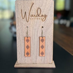 May include: A pair of rectangular, light brown leather earrings with a geometric pattern, displayed on a wooden stand. The stand has the text "Wardog Customs" engraved on it.