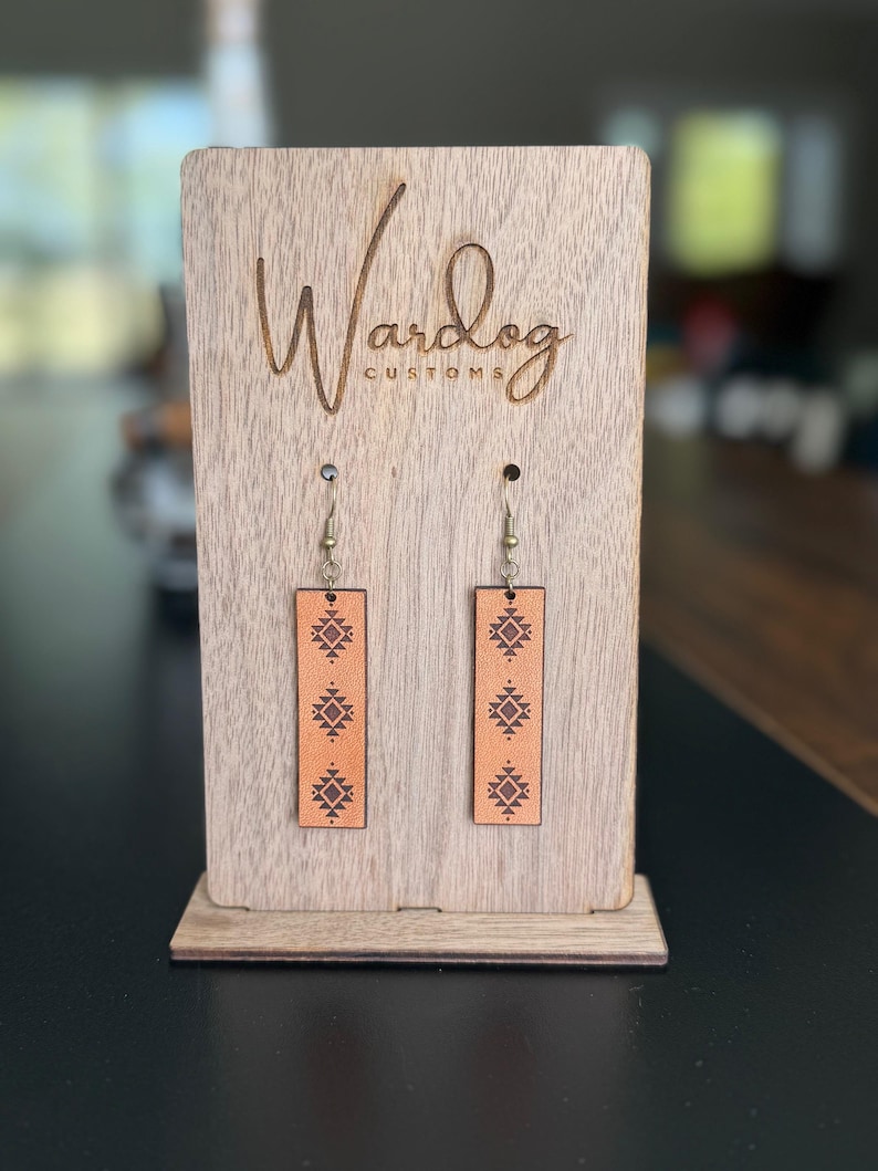May include: A pair of rectangular, light brown leather earrings with a geometric pattern, displayed on a wooden stand. The stand has the text "Wardog Customs" engraved on it.