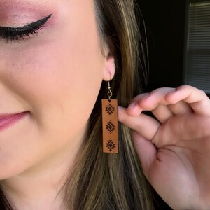 May include: Brown leather earrings with a rectangular shape and a decorative geometric pattern. The earrings are hanging from gold-colored hooks. The earrings are being held up by a hand.