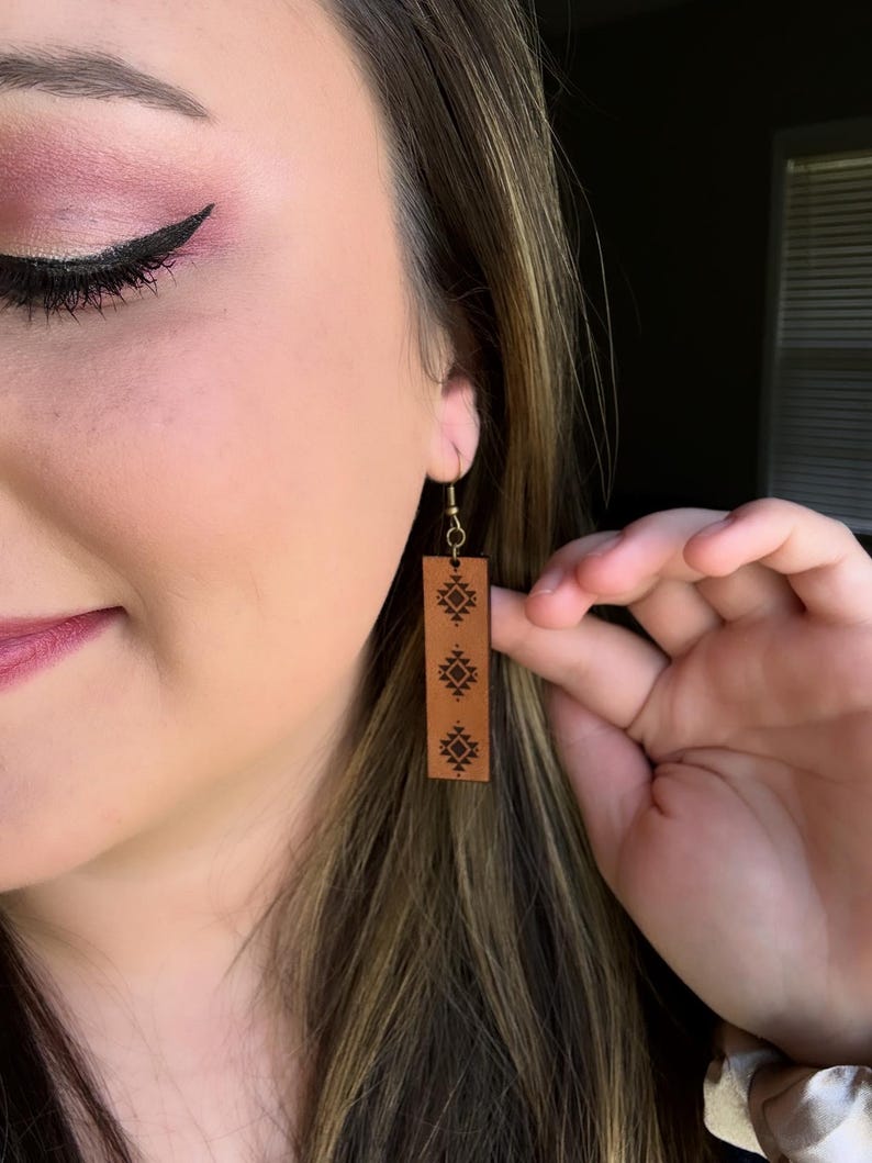 May include: Brown leather earrings with a rectangular shape and a decorative geometric pattern. The earrings are hanging from gold-colored hooks. The earrings are being held up by a hand.